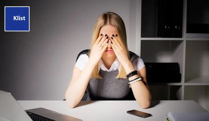 Mulher jovem, loira, sentada num escritório com as duas mãos no rosto, com expressão cansada mostrando os impactos que as 5 piores formas de organizar tarefas causam nas pessoas